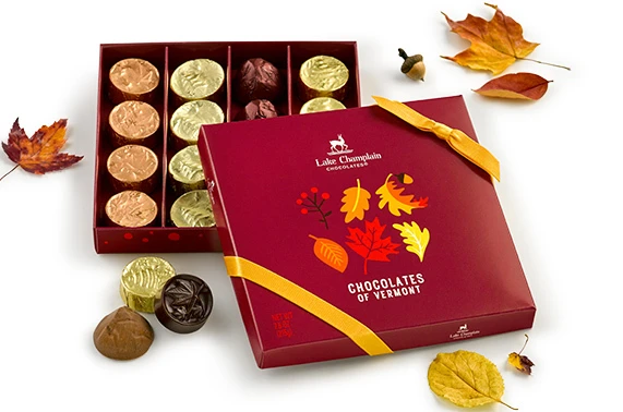 Assorted Chocolates of Vermont in a dark red gift box with red, orange and yellow leaves on it, wrapped and unwrapped chocolates around the gift box with some leaves.