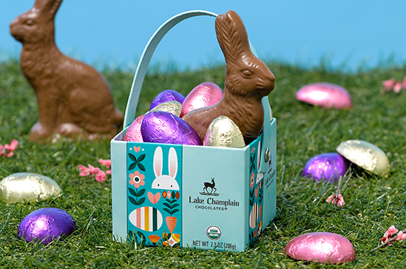 Assorted chocolate eggs wrapped in foils in a cardboard Easter themed gift box with a milk chocolate bunny, with wrapped eggs and a milk chocolate bunny in the background.