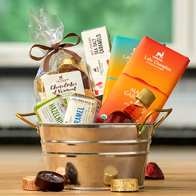 Assorted gourmet chocolates made in Vermont  in a metal gift tin with copper trim sitting on a wooden table top with a maple crunch Chocolates of Vermont wrapped chocolate next to the tin.