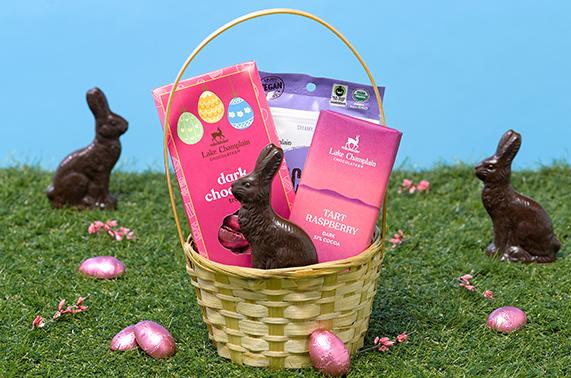 Woven Easter basket filled with a dark chooclate bunny, dark chocolate truffle eggs, a Tart Raspberry chocolate bar and Dark Peanut Butter snack bites, sitting on grass with pink flowers, dark truffle eggs wrapped in pink foils and dakr chocolate bunnies in the background and around the basket.
