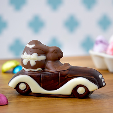 Milk chocolate bunny driving a car "cruisin bunny", sitting on a wooden table with colored chocolate eggs around the bunny in the backgound.