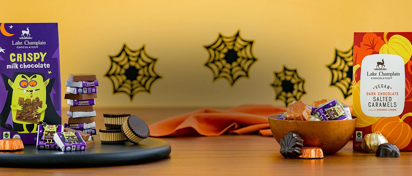 Cripsy milk chocolate Halloween squares box sitting on a black, round, board with wrapped Halloween squares, dark peanut butter chocolates and a wrapped maple leaf chocolate all on the left of the image. On the right is a bix of Vegan Caramel Pumpkins with a bowl of assorted Halloween chocolates and some wrapped and unwrapped chocolates in front of the box.