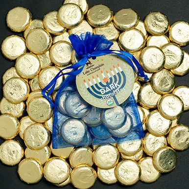 Dark chocolate coins wrapped in gold foil in a giant pile with a royal blue organza bag filled with dark chocolates coins and a Hanukkah themed gift tag, sitting on the pile of coins.