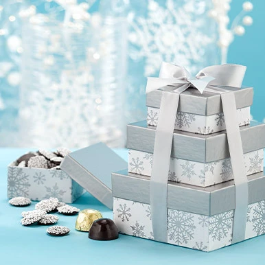 Dark Chocolate Tower of Treasures in silver and white snowflack designed boxes with silver ribbon and a small snowflake designed box on the side filled with dark chocolate nonpareils and nonpareils and wrapped and unwrapped dark chooclate salted caramel jubilees on the left side of the gift box.