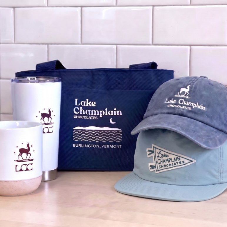 Lake Champlain Chocolates merchandise including a white ceramic mug, white tumbler, blue cooler tote, blue baseball hat and green surf hat all sitting on a wooden surface with white brick in the background.