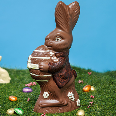 Giant milk chocolate "Goodtime" bunny sitting on grass with colorful foil wrapped chocolate eggs around the bunnt with a blur sky in the background.