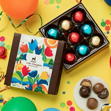 Assorted milk and adark chocolate jubilees in a colorful gift box sitting ona bright yellow background with confetti and balloons.