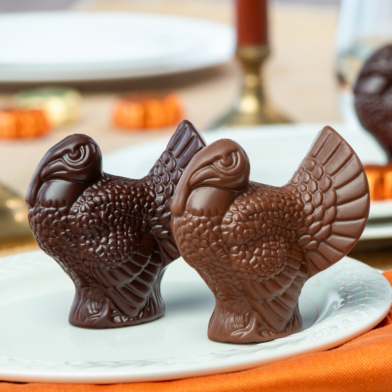 dark and milk chocolate placesetting turkeys sitting on a white ceramic plate on an orange table cloth, with a gold candle holder and a red taper candle in the background alonf with other white ceramic plates and assorted fall chocolates.