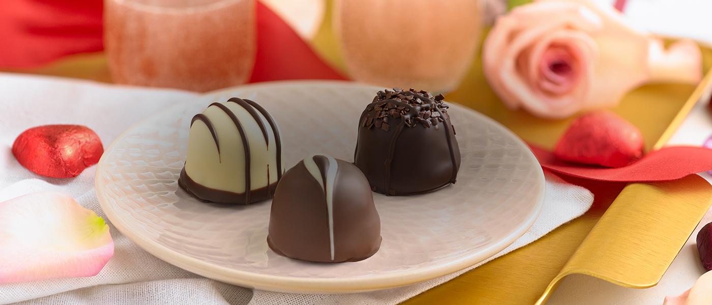 Assorted gourmet chocolate truffles on a  round, white caeramic plate with a gift box in the background and a pink rose and two champagne glasses in the background.
