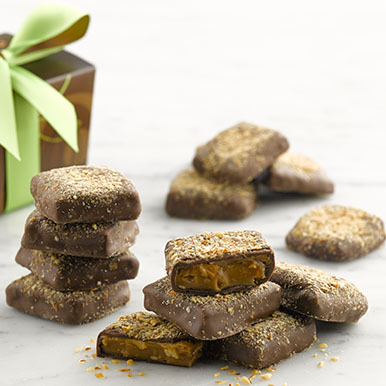 Almond butter crunch pieces stacked ion top of each other on a white marble counter with a brown ballotin box and with a green ribbon in the background.