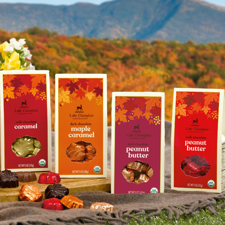 Assorted milk and dark chocolate leaves filled with peanut butter, salted caramel and maple caramel, in fall colored gift boxes, sitting on a wood surface and on a brown blanket with fall colored trees and a mountain in the background and wrapped and unwrapped chocolates in front of the gift boxes.