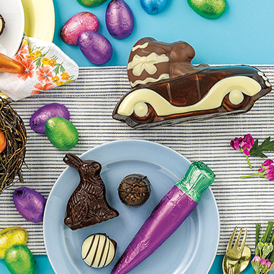 Assorted Easter bunnies, milk and dark chocolate eggs wrapped in colorful foils and a dark chocolate carrot wrapped in purple foil scattered around on a blue background with a black and white striped table cloth.