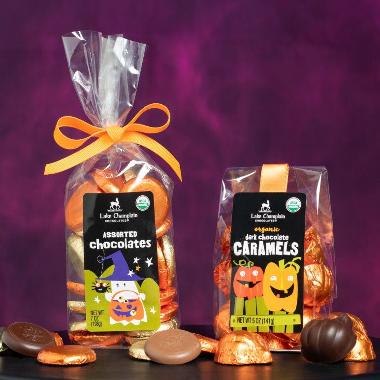 Assorted milk and dark chocolate halloween coins and dark chocolate caramel pumpkins in clear gift bags with orange ribbons and wrapped and unwrapped chocolates around the gift bags.
