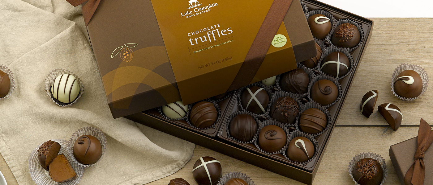 Assorted milk and dark gourmet chocolate truffles in a brown gift box and on a tan colored table cloth on a wood surface.