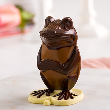 Milk chocolate frog with dark chocolate accents sitting on a white chocolate lily pad with it's arms crossed.