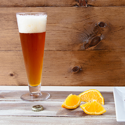 A tall glass of beer sitting on a wooden table with slices of oranges on the side.