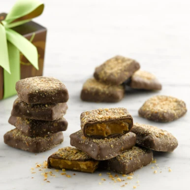 Pieces of Almond Butter Crunch stacked on top of each other making a pile and a tower with a brown gift box tied with a green ribbon in the background.