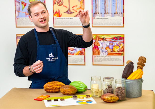 Man wearing a blue apron with the Lake Champlain Chocolates logo on it, holding up a piece of chocolate and talking, standing in front of a table with cocoa pods, jars of cocoa beans, cocoa butter and assorted chocolates in front of him.