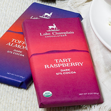 Tart Raspberry Dark Chocolate Bar laying on top of a Toffee & Almonds Dark Chocolate Bar on top of a white fabric table cloth next to a white plate.