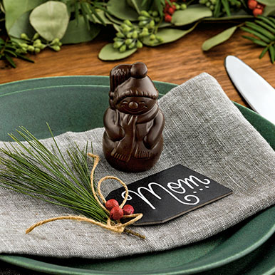 Dark chocolate placesetting snowman on a green ceramic plate with a bunched group of pine needles tired with twine and some berries, with a black name tag that says Mom written in white. 