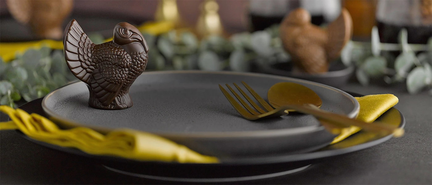Dark chocolate turkey sitting on a dark gray plate with a golden yellow napkin under the palte and a gold fork and spoon on the plate and a milk chooclate turkey with greenery in the background.