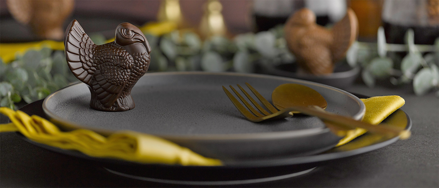 Dark chocolate placesetting turkey on a dark gray ceramic plate with gold colored utensils on the plate and the plate is stiing on a yellow-gold napkin on top of a bigger dark gray ceramic plate with greenery and a milk chocolate turkey in the background.