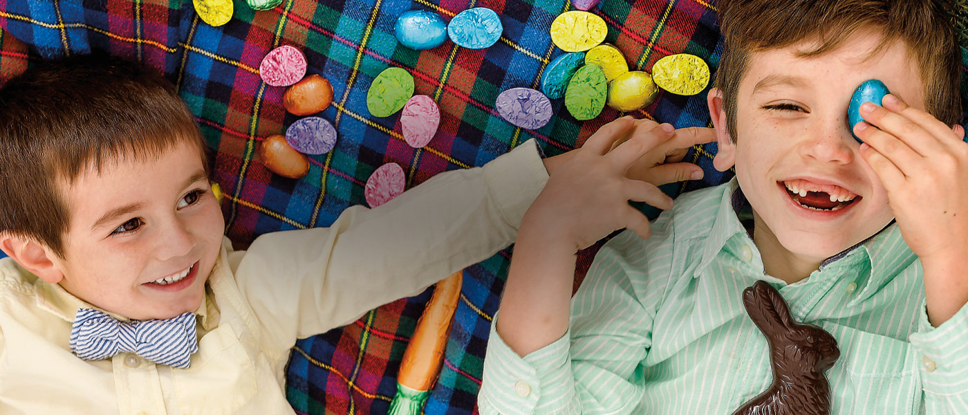 Assorted gourmet Easter chocolates on a black and white stripped table cloth and chocolate eggs wrapped in colorful foils inside a nest, on top of a light blue background.