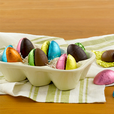 Assorted chocolate eggs wrapped in colorful foils in a ceramic egg carton on a off white table loth with light green stripes.