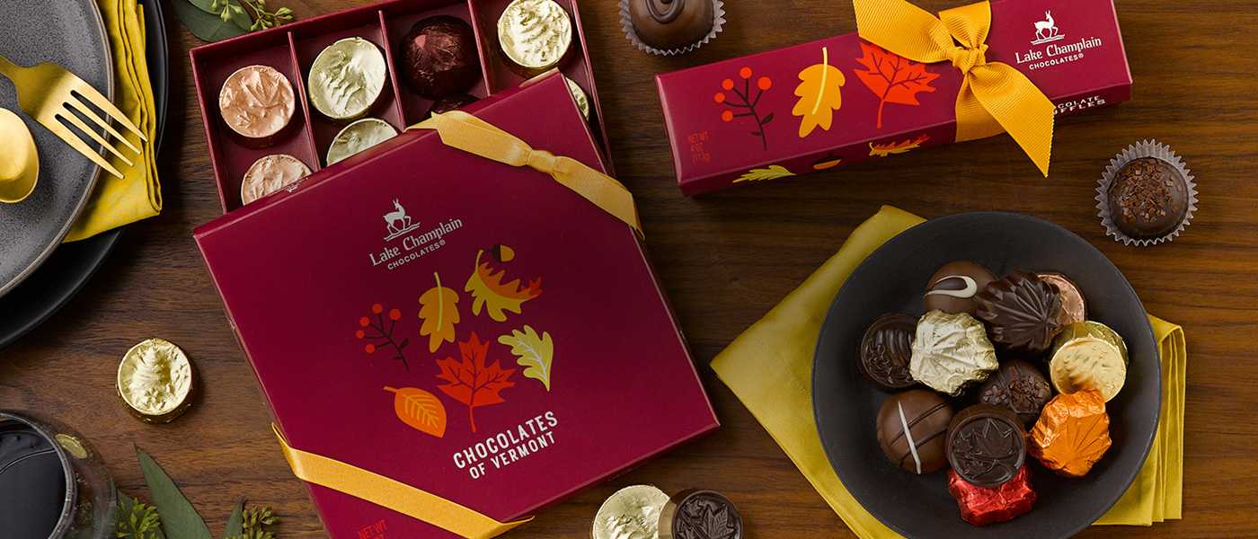 Assorted chocolates in all themed packaging on a wooden surface with a black matte dish filled with assorted chocolates sitting on a gold colored cloth napkin, next to the gift boxes.