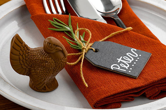 Milk chocolate turkey sitting on a whte ceramic dinner plate with a rustic, burnt orange colored napkin holding a silver fork, knife and spoon with a black name tag tht says "Peter" on it, with twine coming out of the top of the name tag and tied in a bow to a piece of rosemary.
