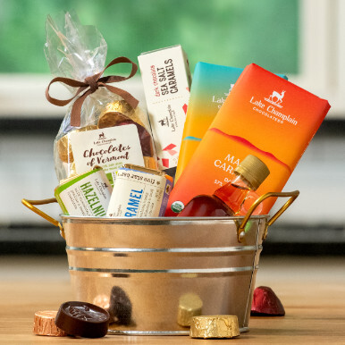 Assorted gourmet chocolates with a small bottle of Vermont maple syrup in a decorative tin bucket with assorted wrapped and unwrapped chocolates around the tin.