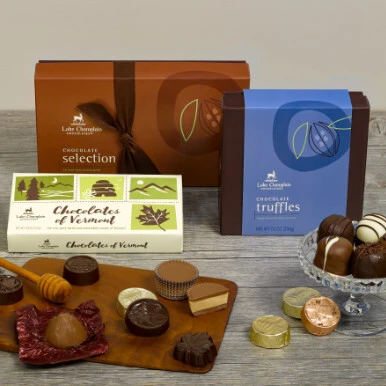 Assorted gourmet chocolates sampler with a 15pc selection gift box, 9pc truffle gift box and 8pc Chocolates of Vermont gift box with wrapped and unwrapped chocolates sitting on a wooden cutting board in front of the boxes and sitting in a glass candy dish.