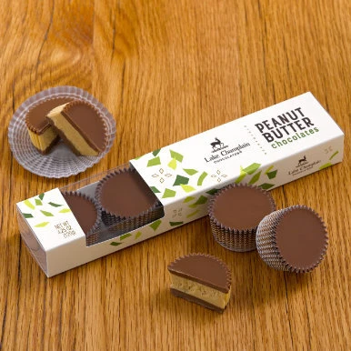 Milk chocolate peanut butter cups sitting on a wooden table and packaged in a gift sleeve with a clear section looking into the gift box and peanut butter cups sitting in front of the gift box..