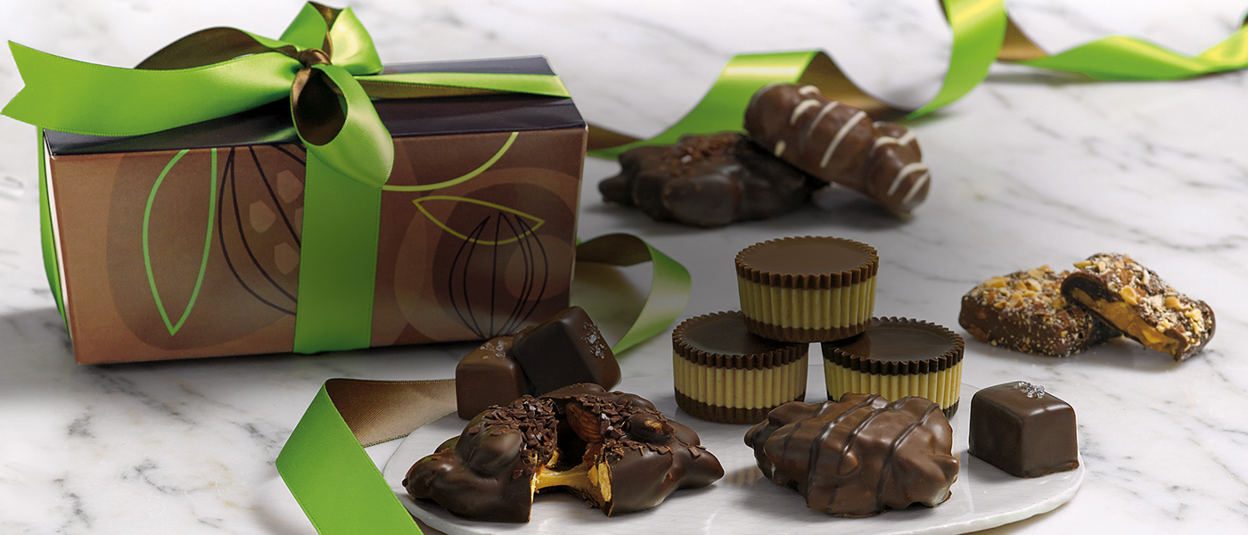 Assorted handcrafted chocolates on a white marble table with a brown ballotin box and a green and brown ribbon in the back.