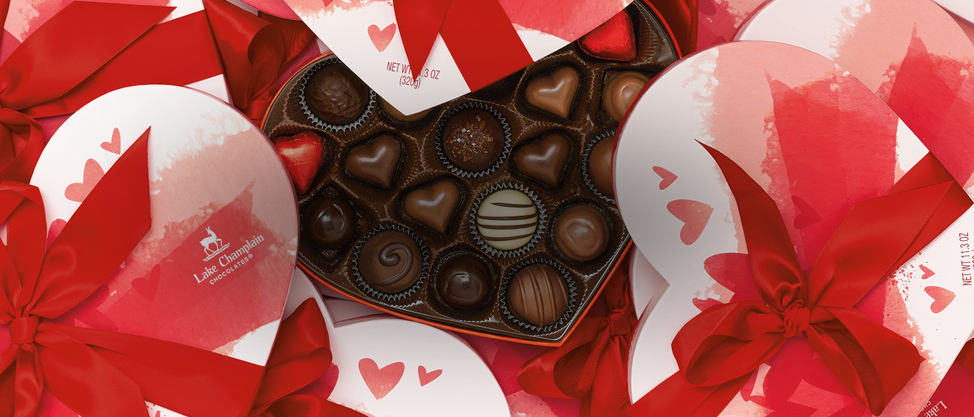 Assorted gourmet chocolates in a heart-shaped box with the lid off of the box and other heart-shaped boxes around the opened box of chocolates.