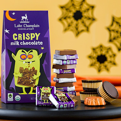Crispy Halloween Milk Chocolate Squares gift box sitting on a black, round, wooden plate with a stack if wrapped and unwrapped chocolate Halloween squares and dark chocolate peanut butter chocolares and a maple caramel leaf on the side and black spider web decorations in the background.