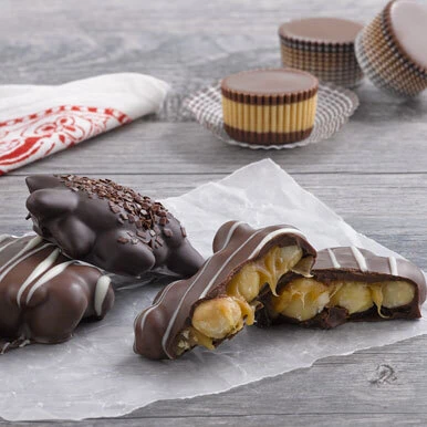 Assorted milk and dark chocolate caramel nut clusters sitting on wax paper on a gray wooden surface with milk chocolate peanut butter cups in the background and a white and red napkin.