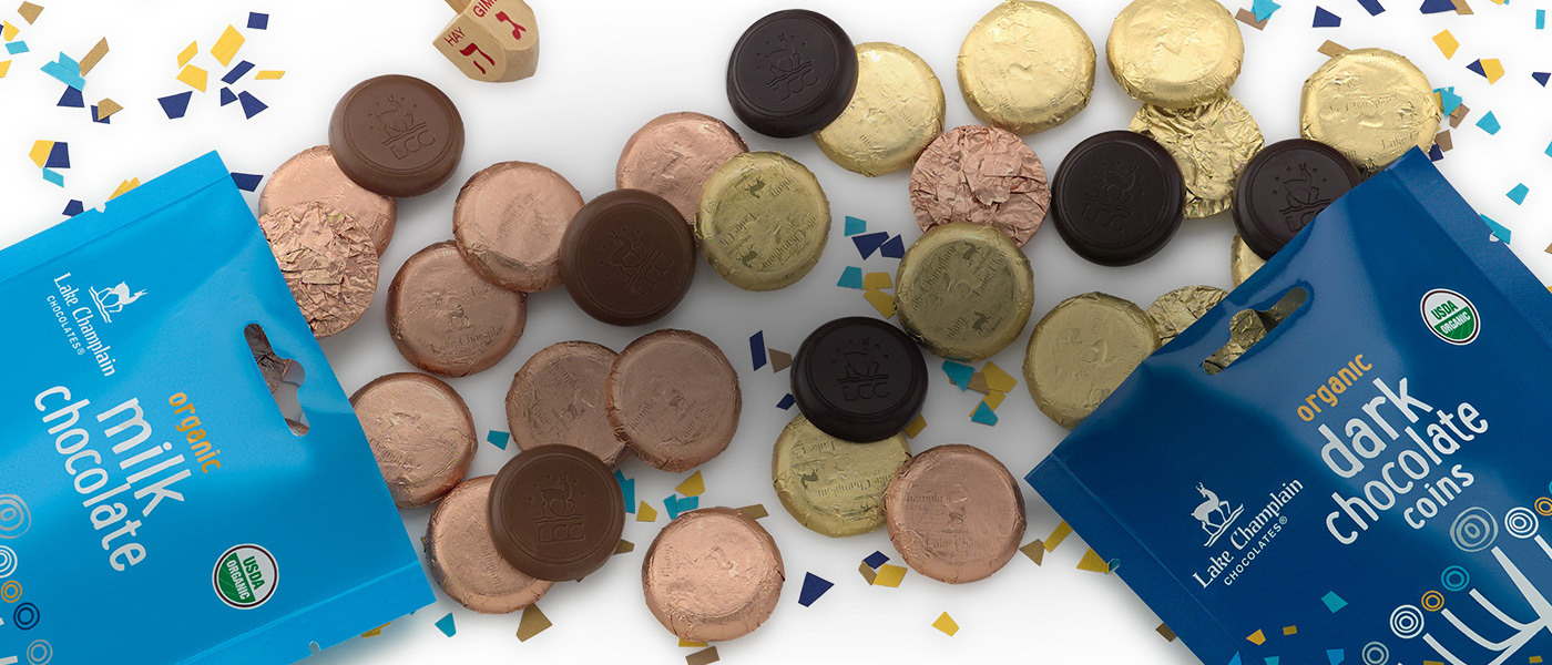 Milk chocolate Hanukkah coins wrapped in copper colored foil, spilling from a light blue Hanukkah themed gift bag and dark chocolate coins wrapped in gold foil pilling out of a dark blue Hanukkah themed gift bag, mixing with the milk chocolate coins, with different shared of blue, yellow and gold confetti in the background.