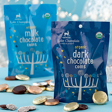 Dark blue dark chocolate hannukah coins bag infront of a light blue milk chocolate hanukkah coins bag with assorted chocolate coins and nonpareils around the bags.