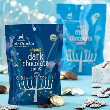 Dark chocolate Hanukkah coins in a dark blue Hanukkah themed gift bag with assorted wrapped and unwrapped coins around the bag and dark chocolate nonpareils around the bag and a light blue milk chocolate Hanukkah coin bag in the background.