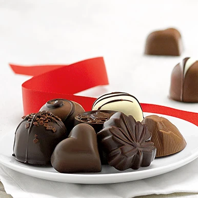 Assorted gourmet chocolates on a round white plate with a red ribbon and unwrapped chocolates in the background.