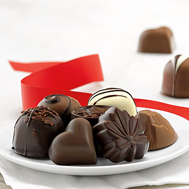 Assorted unwrapped chocolates on a white ceramic plate with a red ribbon and a truffle and a chocolate heart in the background..