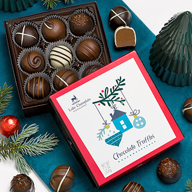 Assorted gourmet chocolate truffles in a holiday theme gift box on a dark teal table cloth with truffles around the gift box and a green ceramic tree next to the box.
