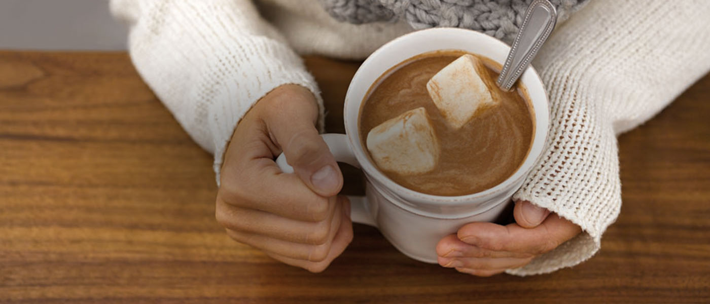 Woman wearing a white knitted sweater and a gray scarf holding a white ceramic mug of hot chocolate with two large marshmallows in it.