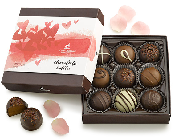 Assorted gourmet chocolates in a holiday themed gift box sitting on greenery with berries and white Christmas lights.