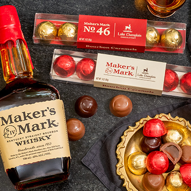 Milk and dark chocolate Maker's Mark Bourbon caramels in clear gift boxes with wrapped and unwrapped caramels in a gold dish and a bottle of Maker's Mark Whisky next to the chocolates.