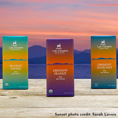 Sea Salt Toffee, Crunchy Peanut, and Crunchy Hazelnut chocolate bars standing straight up on a wooden table with a sunset and mountain view in the background. Sunset photo credit: Sarah Lavoie.