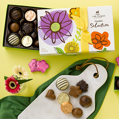 Assorted gourmet chocolates in a spring themed gift box with vegan chocolate flowers wrapped in pink foils and assorted gourmet chocolates sitting on a white cutting board.