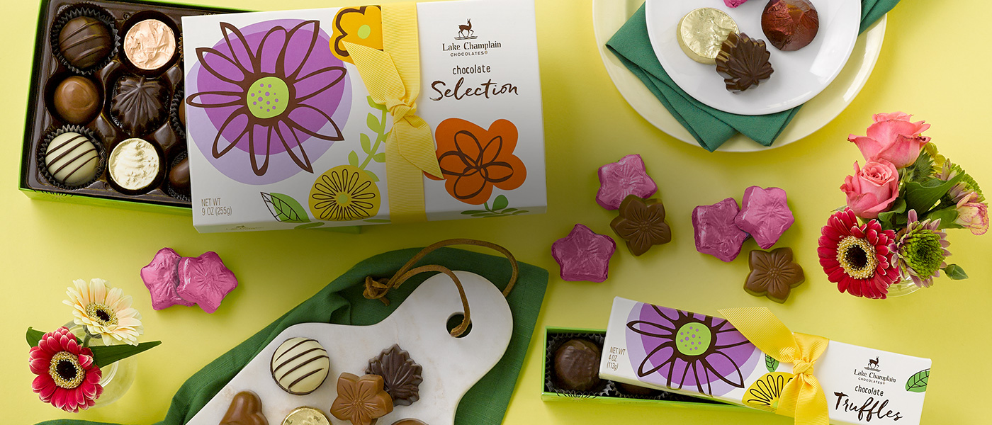 Assorted gourmet chocolates in spring themed gift boxes with dark chocolate vegan caramel flowers wrapped in pink foils and a variety of colorful flowers in vases, on a yellow background.