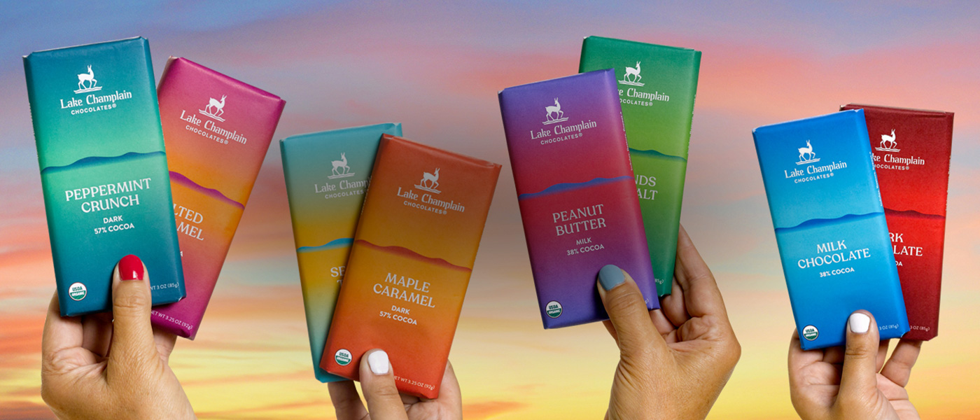 Assorted milk and dark chocolate bars in colorful packagin being helod up by hands with colorful painted nails, against a sunset background.